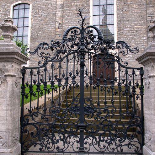 Schwarze, kunstvoll verzierte Eisen-Türen führen zu Steinstufen vor einer grauen Wand mit großen Fenstern.