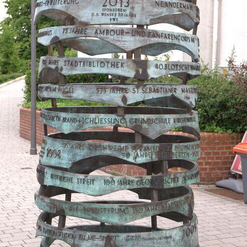 Skulptur aus gebogenem Metall mit diversen gravierten Jahreszahlen und Ereignissen im öffentlichen Raum.