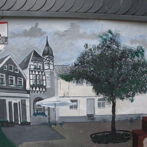 Wandgemälde eines historischen Gebäudes mit Fachwerk-Architektur und einem Baum auf einer gepflasterten Fläche.