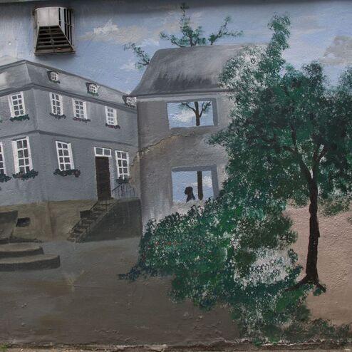 Wandgemälde von Gebäuden, einem Brunnen und Bäumen in einer städtischen Umgebung. Fenster zeigen Umrisse von Personen.