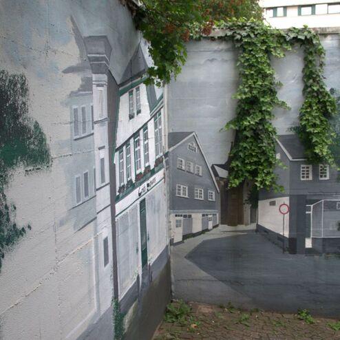 Wandgemälde eines Stadtelements mit Gebäuden und einem gepflasterten Weg, umgeben von grüner Vegetation.