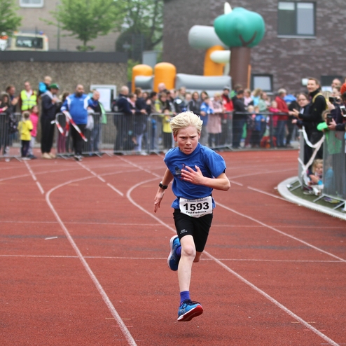 Ein Junge mit blonden Haaren sprintet auf einer Laufbahn, während Zuschauer auf der Tribüne jubeln.