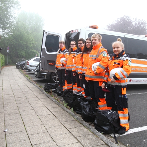 Gruppe junger Rettungskräfte in oranger Uniform mit Helmen steht neben einem Einsatzfahrzeug auf einem Gehweg.