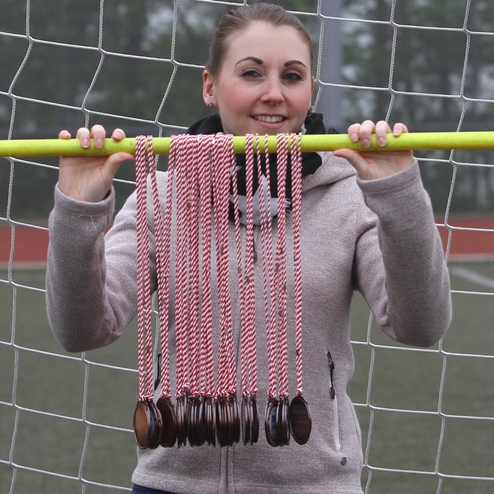 Frau hält eine gelbe Stange mit mehreren Medaillen und roten Bändern vor einem Fußballtor auf einem Sportplatz.