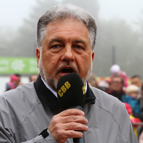Ein Mann mit grauen Haaren hält ein Mikrofon und spricht vor einer Menschenmenge in nebliger Umgebung.
