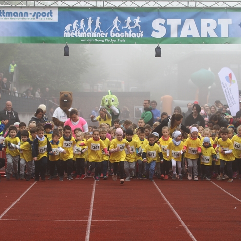 Start der Kinderläufe mit zahlreichen kleinen Teilnehmern in gelben T-Shirts auf einer roten Laufbahn, umgeben von jubelnden Zuschauern.
