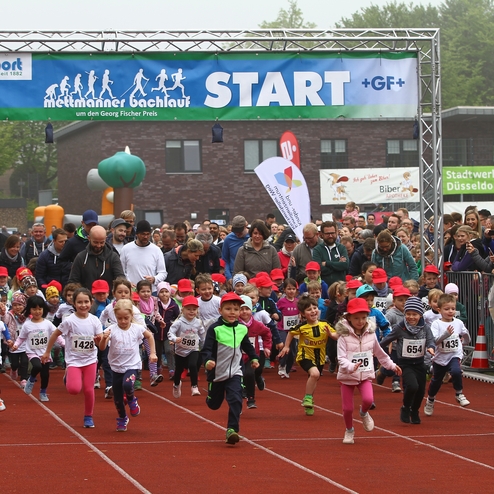 Gruppenauslauf von Kindern in Sportkleidung und roten Caps beim Start eines Laufwettbewerbs, Zuschauer im Hintergrund.
