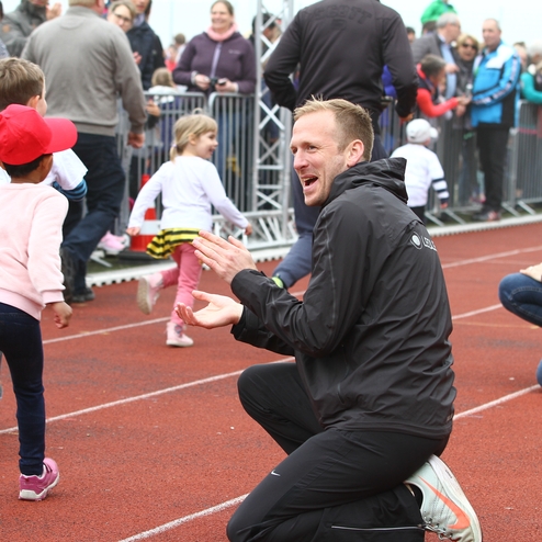 Kinder laufen auf einer Laufbahn, während Erwachsene am Rand anfeuern und sich freuen.