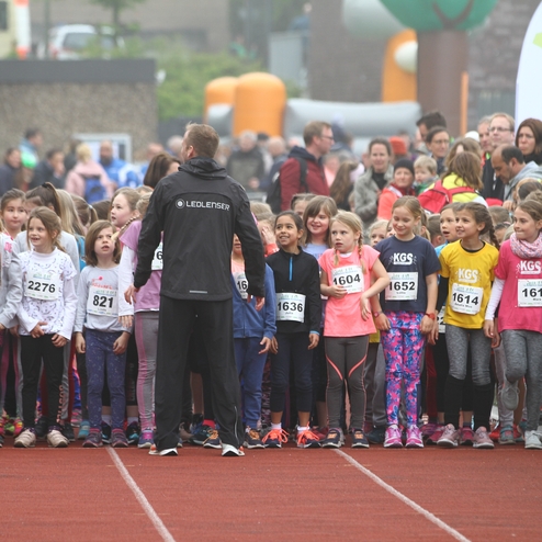 Kinder stehen an der Startlinie eines Wettlaufes, während ein Betreuer sie anweist. Zuschauer sind im Hintergrund sichtbar.