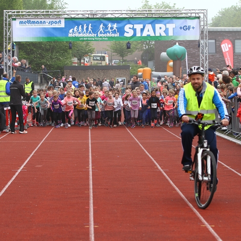 Startschuss eines Kinderlaufs mit zahlreichen Teilnehmern auf der Laufbahn, im Hintergrund jubelnde Zuschauer und ein Maskottchen.