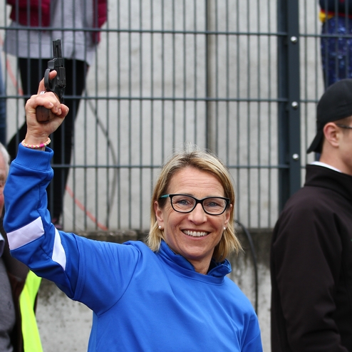 Lächelnde Frau in blauer Jacke hält eine Startpistole hoch, im Hintergrund ein älterer Mann und ein junger Mann.