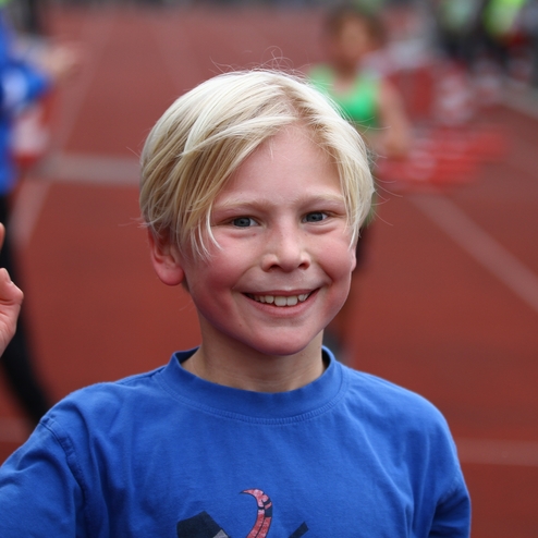 Lächelnder Junge mit blonden Haaren winkt auf einer Laufbahn während eines Sportevents.