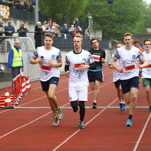 Sechs Jugendliche in Laufkleidung absolvieren einen Wettlauf auf einer roten Laufbahn, Zuschauer beobachten vom Rand.