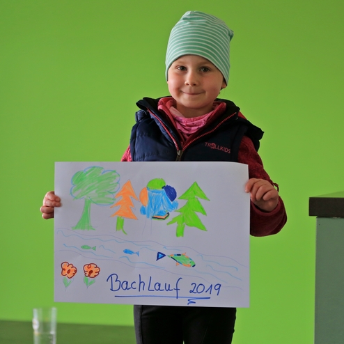 Kind mit Mütze hält ein buntes Plakat mit einer Landschaftszeichnung und der Aufschrift „Bachlauf 2019“, vor grüner Wand.