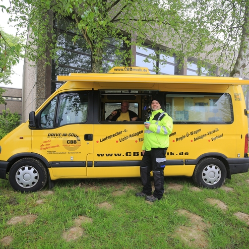 Gelber Transporter mit zwei Männern, einer steht davor, der andere lächelt aus dem Fenster, umgeben von Bäumen und Gras.