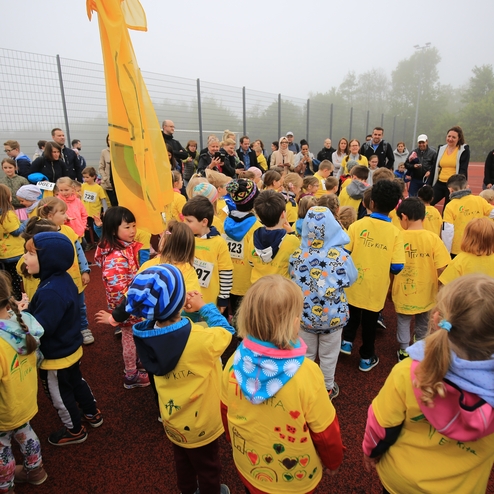 Gruppiertes Aufgebot von Kindern in gelben T-Shirts, umgeben von Zuschauern auf einem Sportplatz im Nebel.