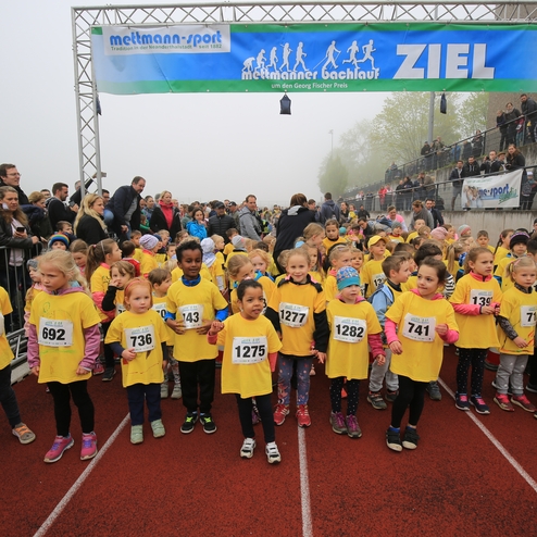 Mehrere Kinder in gelben T-Shirts mit Startnummern stehen auf einer Laufbahn, umgeben von Zuschauern und einer Wolkendecke.