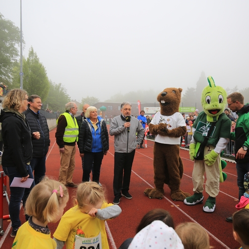 Gruppenszene bei einem Sportereignis mit Sprechern, Maskottchen und Zuschauern auf einer Laufbahn im Nebel.