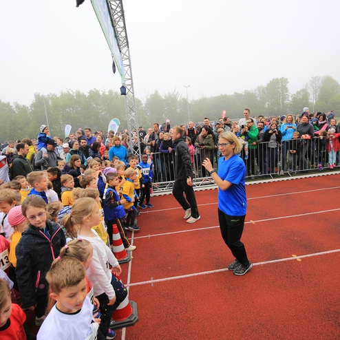 Kinder stehen auf einer Laufbahn, während Erwachsene im Hintergrund zuschauen und ein Event leitet eine Person in blauer Bekleidung.