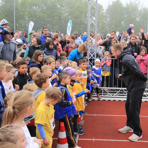 Gruppe von aufgeregten Kindern in Sportbekleidung wartet an der Startlinie eines Wettlaufs, umgeben von Zuschauern.
