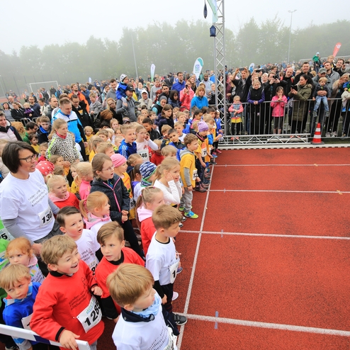 Kinder in bunten T-Shirts stehen an der Startlinie eines Rennens, während eine Menschenmenge sie anlachtet.