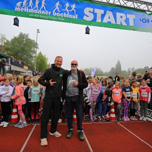 Zwei Männer posieren lächelnd vor einer Startlinie, während eine Gruppe von Kindern für ein Rennen aufgereiht steht.