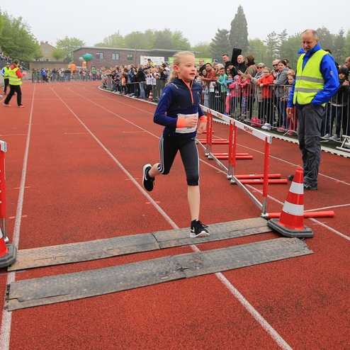 Mädchen läuft beim Hürdenlauf über eine rote Laufbahn, Publikum applaudiert im Hintergrund.