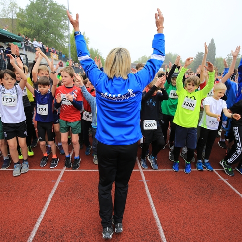 Gruppierung von Kindern in Wettkampfbekleidung, die begeistert zu einer Trainerin mit erhobenen Händen auf einer Laufbahn stehen.