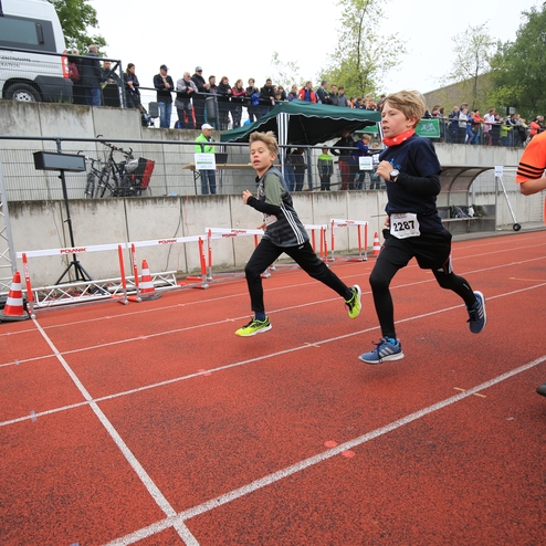 Drei Kinder laufen auf einer roten Laufbahn während eines Wettkampfs, Zuschauer stehen auf einer Tribüne im Hintergrund.