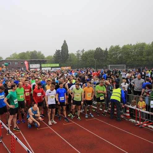 Große Menschenmenge von Läufern auf einem Sportplatz, bereit für den Wettkampf, umgeben von Zuschauern und Gerätschaften.