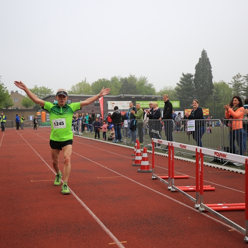 Ein älterer Läufer mit Nummer 1245 überquert die Ziellinie und jubelt, umgeben von applaudierenden Zuschauern.