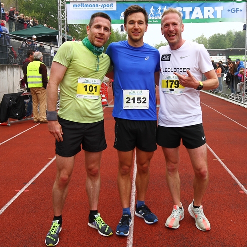 Drei Läufer in Sportkleidung stehen lächelnd an der Startlinie eines Wettbewerbs auf einer roten Laufbahn.