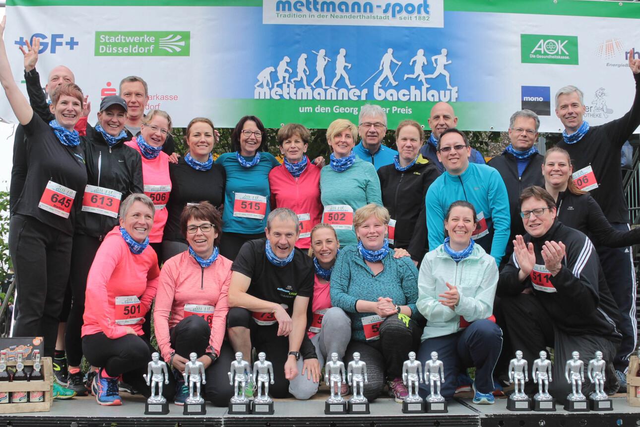 Gruppenselfie von Teilnehmern eines Lauf-Events, die auf einer Bühne vor Pokalen posieren, mit Startnummern auf der Kleidung.
