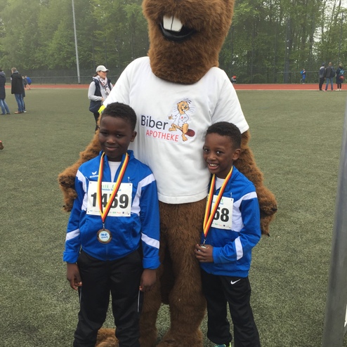 Zwei Kinder mit Medaillen stehen neben einem bärenförmigen Maskottchen auf einem Sportplatz.