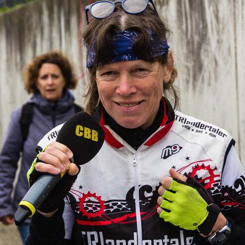 Lächelnde Frau mit Mikrofon, trägt Sportbekleidung und Handschuhe, im Hintergrund eine weitere Person.