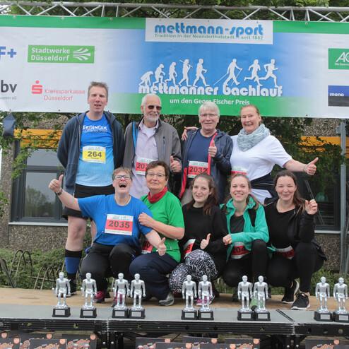Gruppenbild von Teilnehmern des Mettmanner Bachlaufs auf einem Podium vor einer Sponsorenwand mit Pokalen.