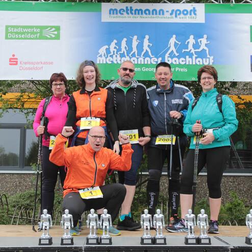 Sechs Teilnehmer posieren mit Pokalen nach dem Mettmann Bachlauf vor einer Sponsorenwand.