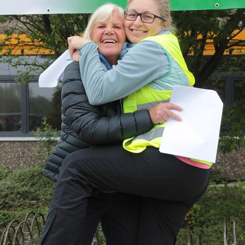 Zwei Frauen umarmen sich lächelnd, eine trägt eine neonfarbene Weste, die andere hält Unterlagen in der Hand.