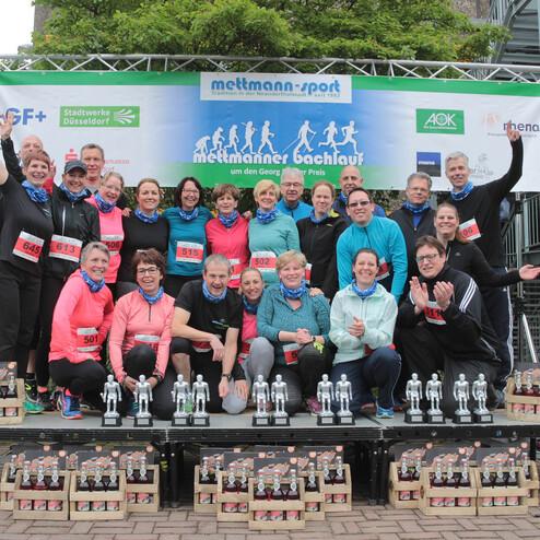 Gruppenselfie von Teilnehmern eines Lauf-Events, die auf einer Bühne vor Pokalen posieren, mit Startnummern auf der Kleidung.