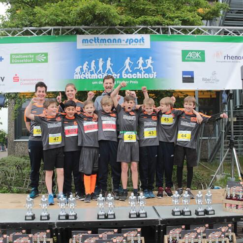 Gruppe von Kindern auf einer Bühne beim Mettmanner Bachlauf, jubelnd mit Medaillen und Pokalen im Vordergrund.