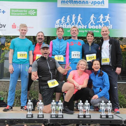 Gruppe von Läufern posiert lächelnd auf einer Bühne mit Pokalen, nach dem Mettmann Bachlauf.