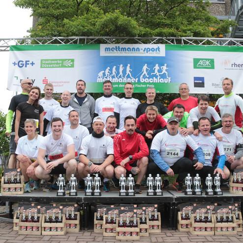 Gruppe von Männern in Sportkleidung posiert auf einer Bühne mit Pokalen und Medaillen nach einem Laufwettbewerb.