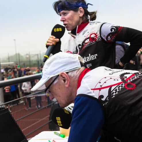 Zwei Personen bei einer Veranstaltung, eine spricht ins Mikrofon, die andere schaut auf einen Laptop und Notizen.