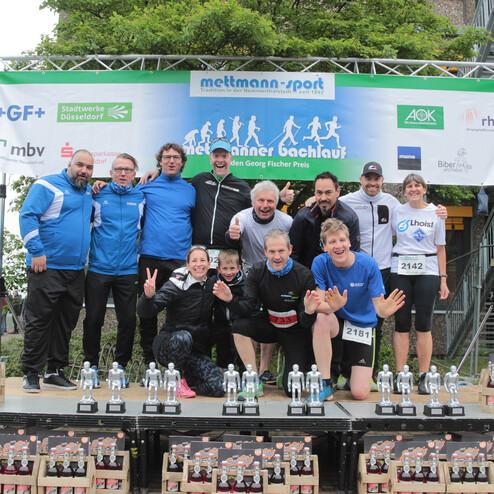 Gruppenselfie von Läufern auf einer Siegerehrungsbühne mit Pokalen vor einem Sportbanner.
