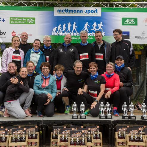 Gruppe von Läufern auf einer Bühne, umgeben von Pokalen und Getränken, während sie an einem Wettlauf teilnehmen.