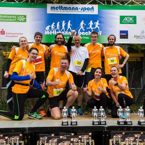 Gruppe von Sportlern in orangefarbenen Shirts posiert lächelnd mit Pokalen nach einem Laufwettbewerb.
