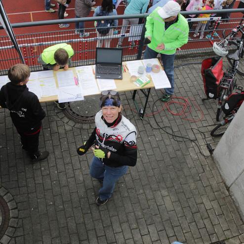 Eine Person in sportlicher Kleidung lächelt und zeigt den Daumen hoch, während im Hintergrund Kinder an einer Laufveranstaltung teilnehmen.