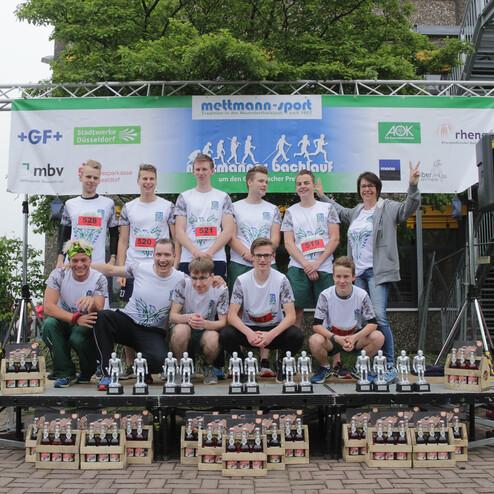Gruppe von Läufern in T-Shirts mit Nummern auf der Siegerehrung, umgeben von Pokalen und Preisen.