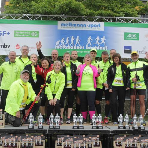 Gruppe von Läufern in neonfarbener Sportbekleidung posiert auf einer Siegerehrung mit Pokalen und Medaillen.
