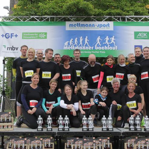 Gruppe von Läufern in schwarzen Shirts mit Startnummern, posierend vor einer Bühne mit Pokalen und Sponsorenbanner.
